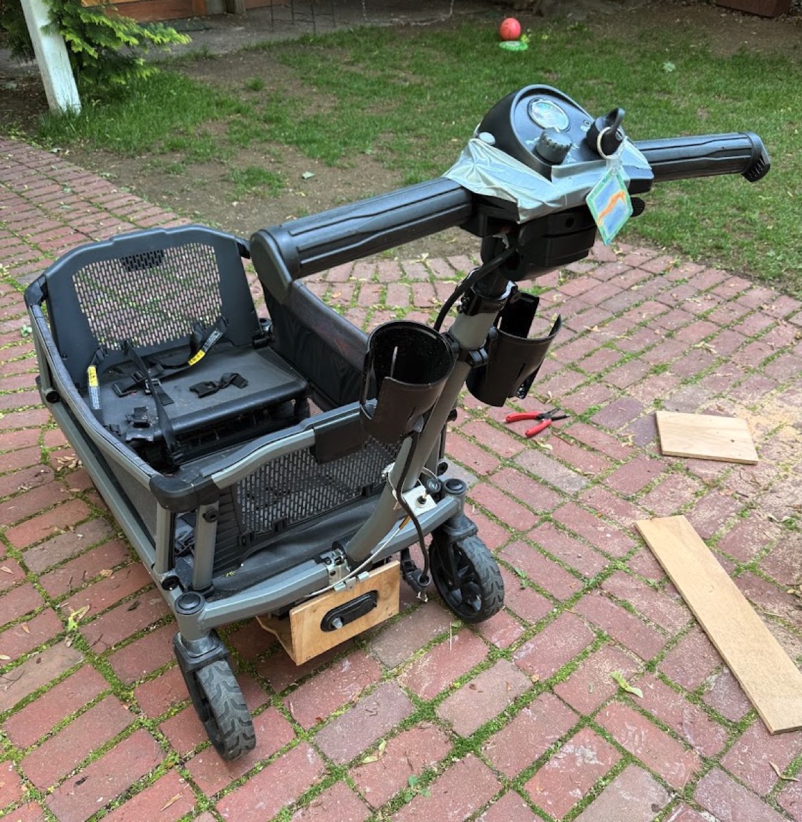 A Veer City Cruiser XL wagon with a mobility-scooter front end grafted on: tiller steering column, handlebar controls, single front wheel, and a plywood battery box mounted under the frame.